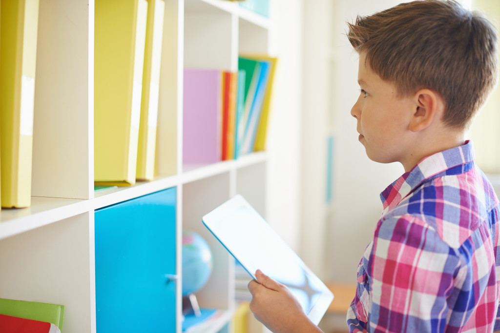 Serious schoolboy with touchpad looking at books on shelf
